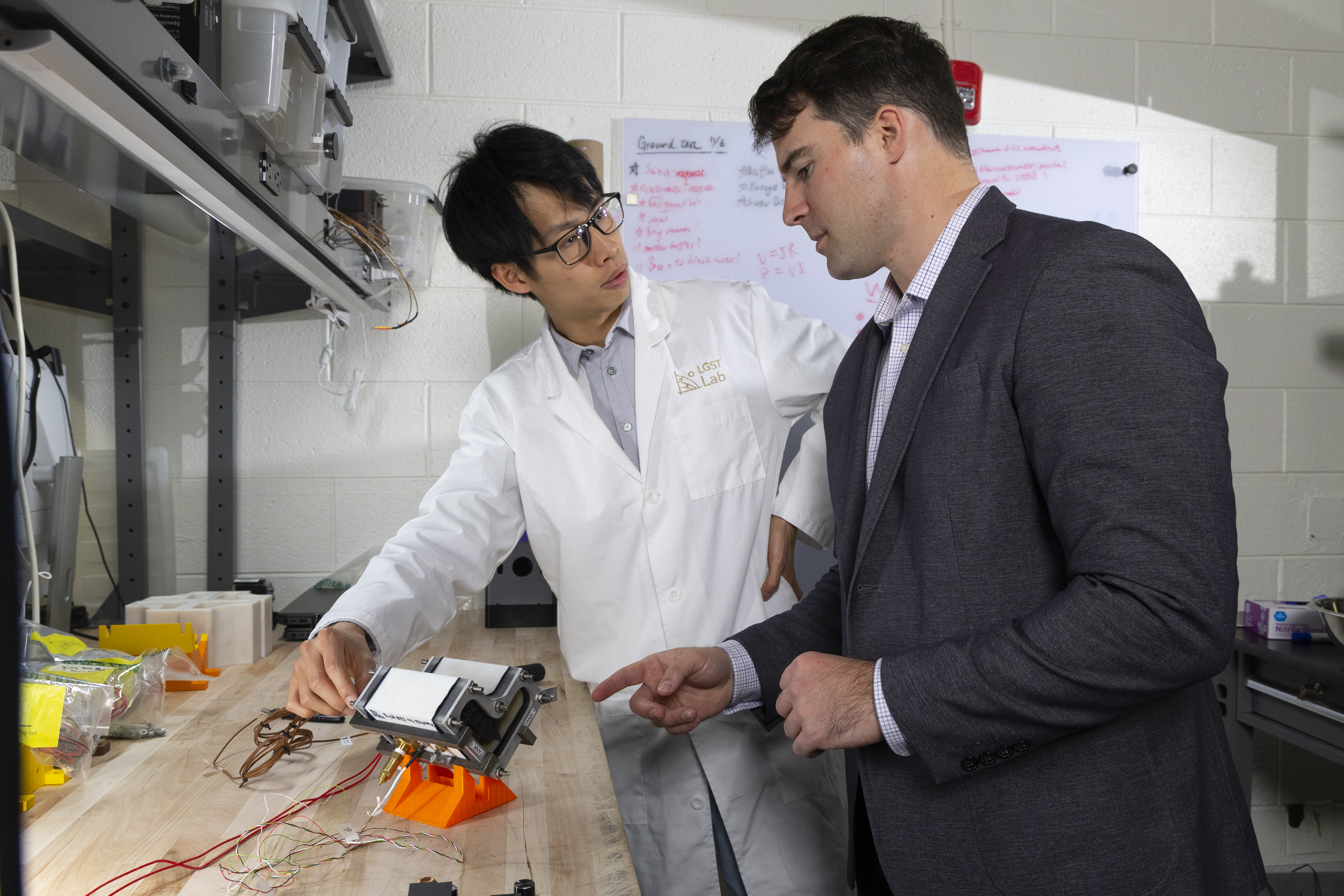 Two researchers examine a small experimental device on a laboratory workbench. One researcher in a white lab coat gestures toward the instrument while the other points to a component. Wires extend from the device, which is mounted on a stand among lab equipment and materials. Whiteboards and shelving with additional research tools are visible in the background.