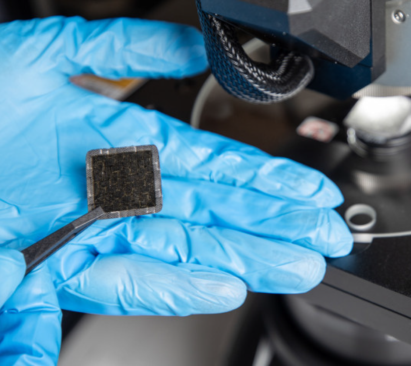 Close up of a hand in blue latex gloves holding a sample of carbon fiber reinforced polymer (CFRP) material with tweezers.